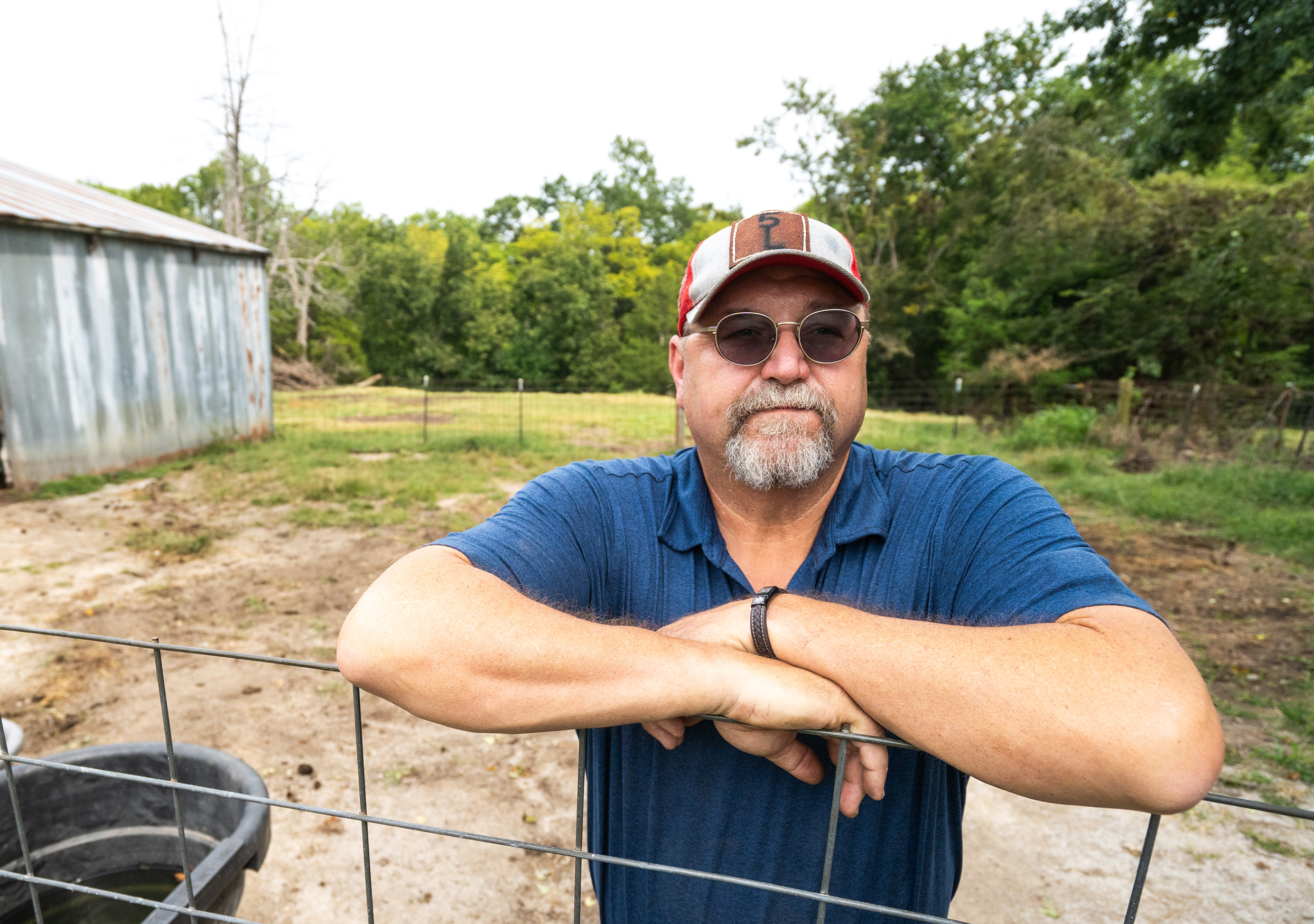 Man wearing round sunglasses and a baseball cap leaning on a wire fence