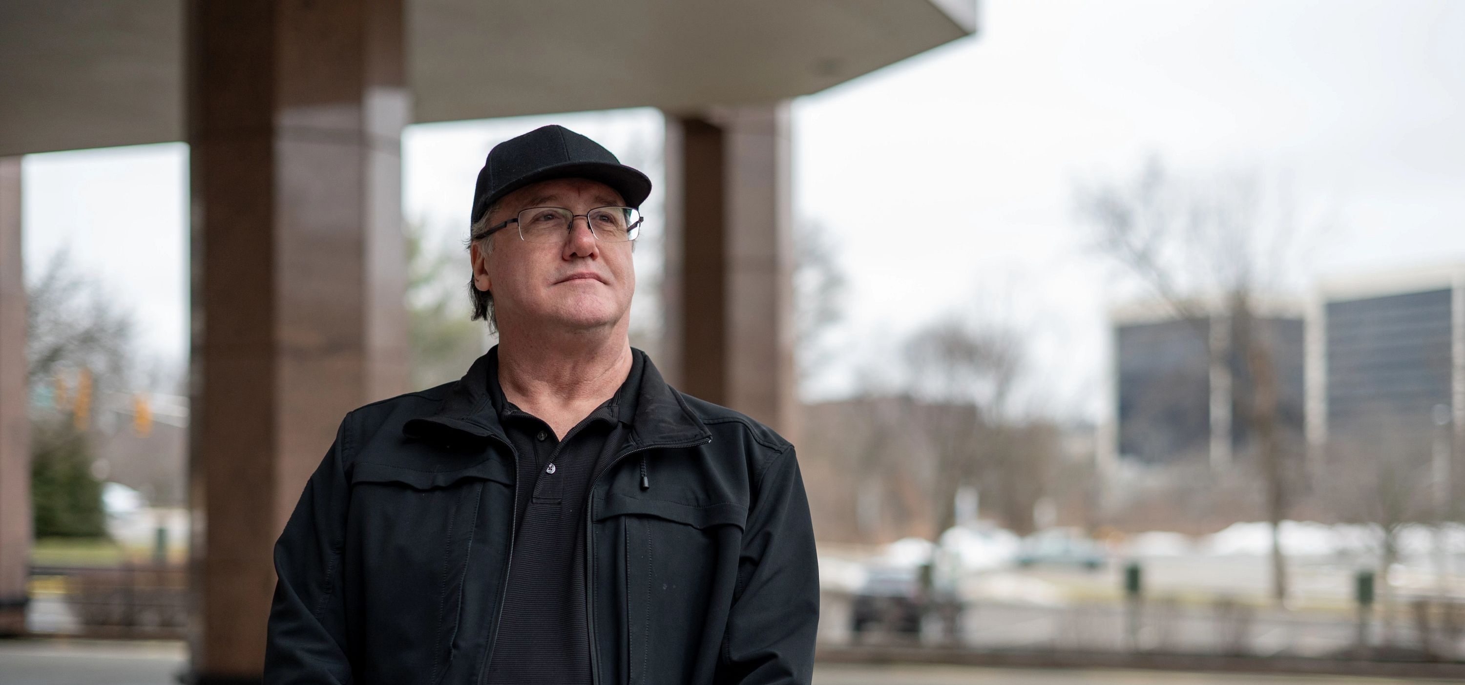 Man wearing a hat and glasses looking up to the right standing in front of a building