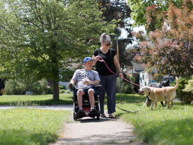 Son in a wheelchair walking down a sidewalk past a stop sign with his mom/caregiver