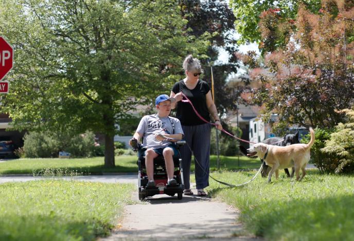 Son in a wheelchair walking down a sidewalk past a stop sign with his mom/caregiver