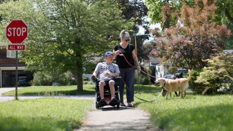 Son in a wheelchair walking down a sidewalk past a stop sign with his mom/caregiver